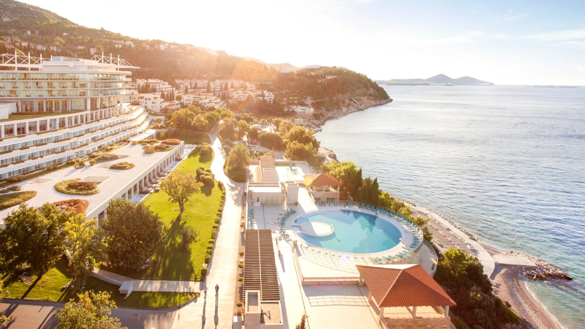 Aerial view of a coastal resort with a circular pool beside the sea at sunset.
